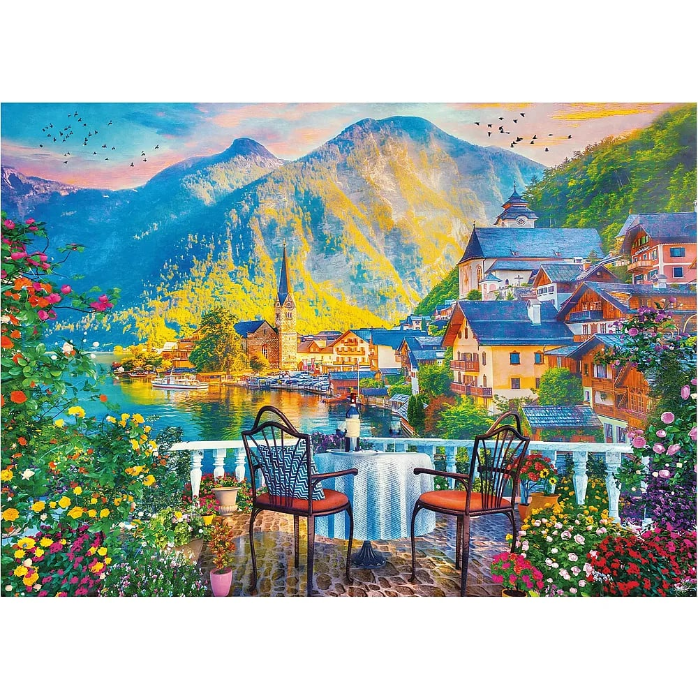 Blick von einem Balkon mit Tisch und Blumen auf einen See, eine Stadt und Berge bei Sonnenuntergang.