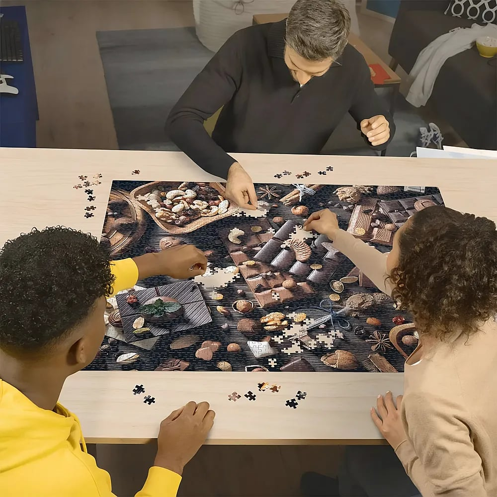 Drei Personen sitzen an einem Tisch und legen ein gemeinsames Puzzle aus, das Schokolade und Nüsse zeigt.