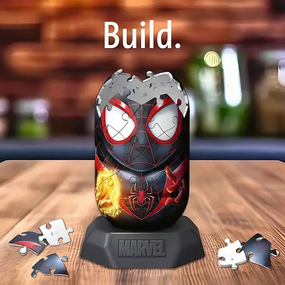 Eine Spider-Man 3D-Puzzlekapsel auf einem Marvel-Ständer mit losen Puzzleteilen auf einem Holztisch und dem Wort "Build".