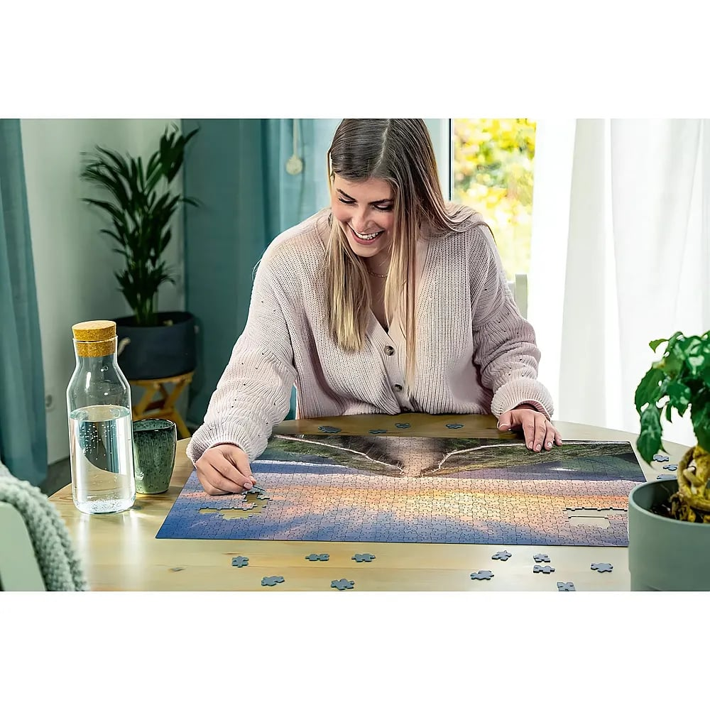 Eine lächelnde Frau in einer rosa Strickjacke sitzt an einem Holztisch und löst ein großes Puzzle.
