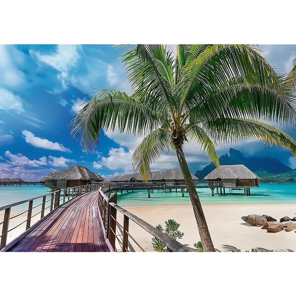 A wooden pier leads to overwater bungalows on clear turquoise water under a blue sky with a palm tree in the foreground.