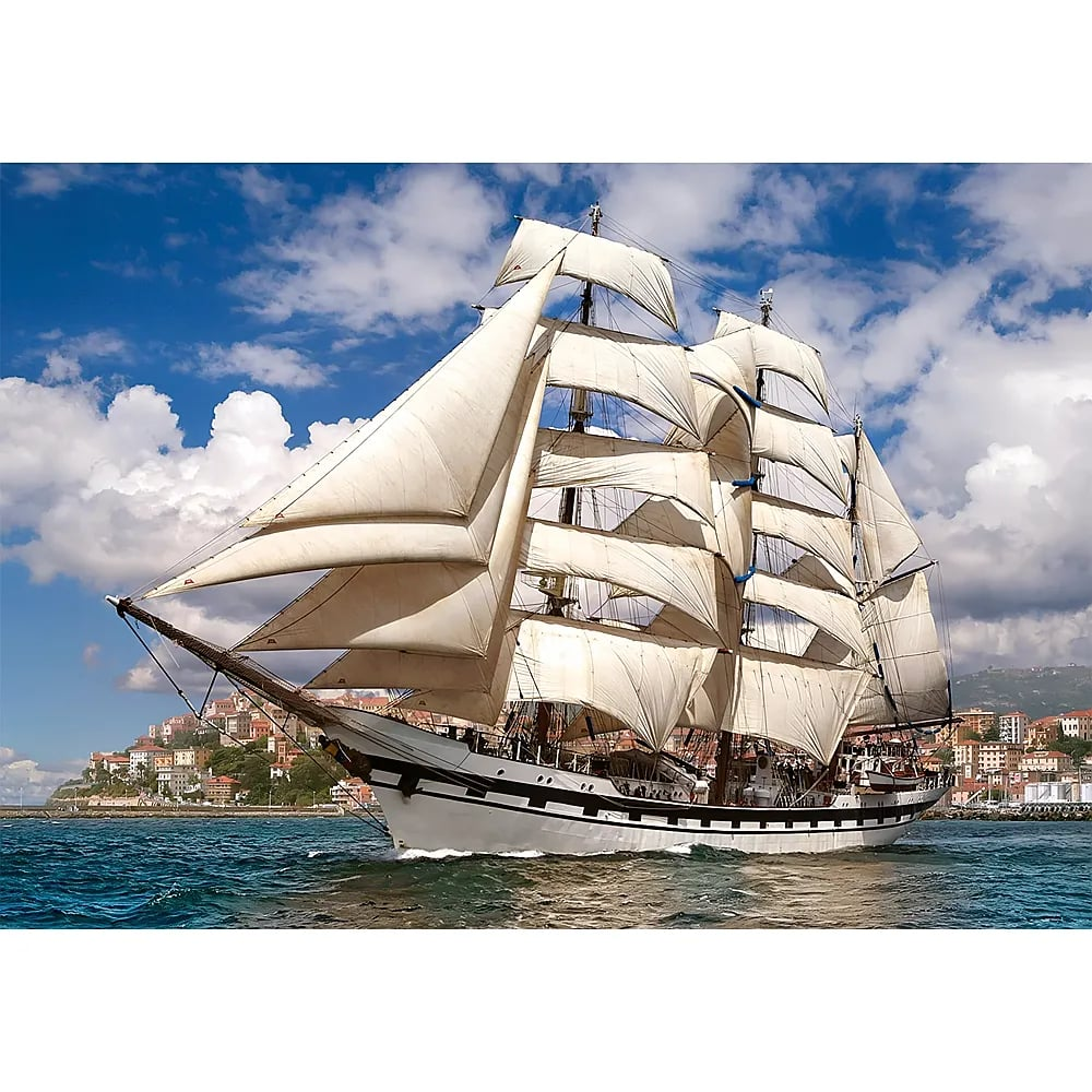 Ein großes, weißes Segelschiff segelt auf blauem Wasser vor einer Küstenstadt unter einem bewölkten Himmel.