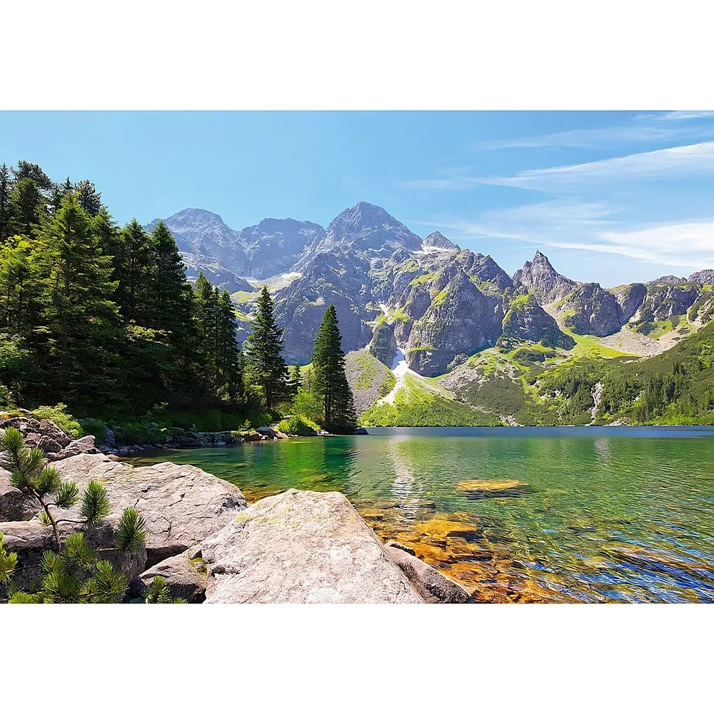 Lac alpin clair avec de grands rochers au premier plan, des pins verts et des montagnes majestueuses en arrière-plan.