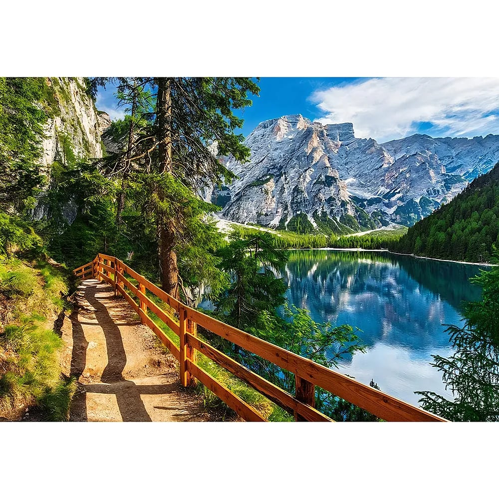 Ein hölzerner Wanderweg führt an einem blauen See vorbei, im Hintergrund erheben sich schneebedeckte Berge.