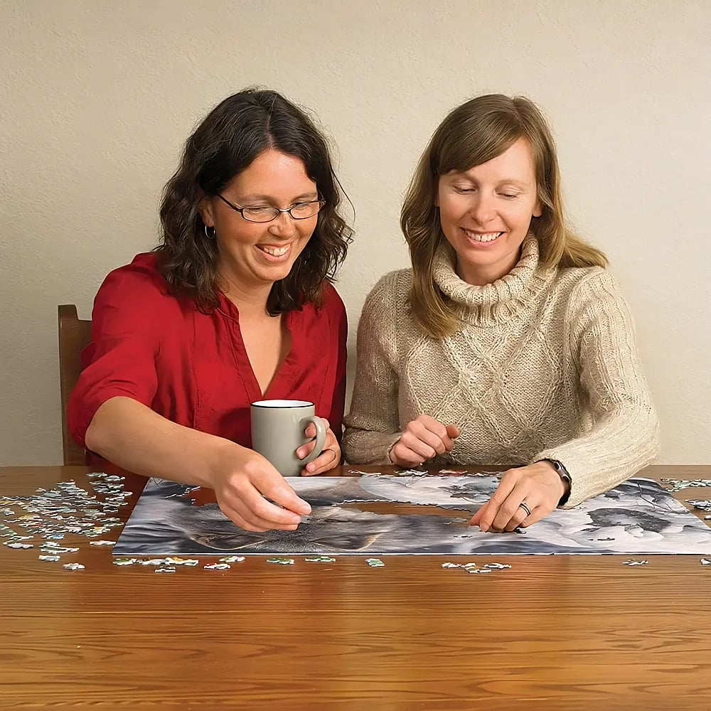 Zwei Frauen lächeln an einem Holztisch und setzen gemeinsam ein großes Puzzle zusammen.