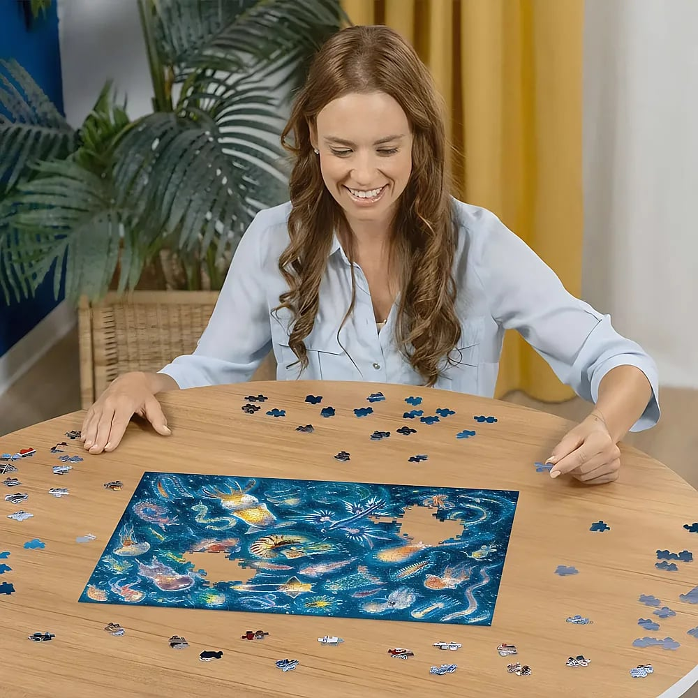 Eine lächelnde Frau sitzt an einem runden Holztisch und macht ein ozeanblaues Puzzle.
