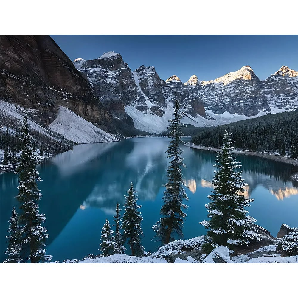 Schneebedeckte Berge und Immergrüne spiegeln sich unter blauem Himmel in einem ruhigen türkisen Alpensee.