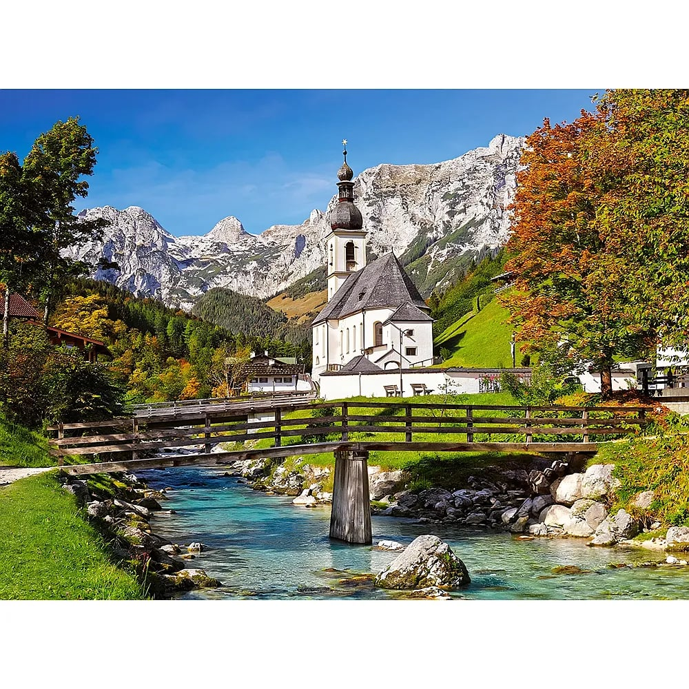 Eine Holzbrücke führt über einen klaren Bach zu einer weißen Kirche vor felsigen Bergen.