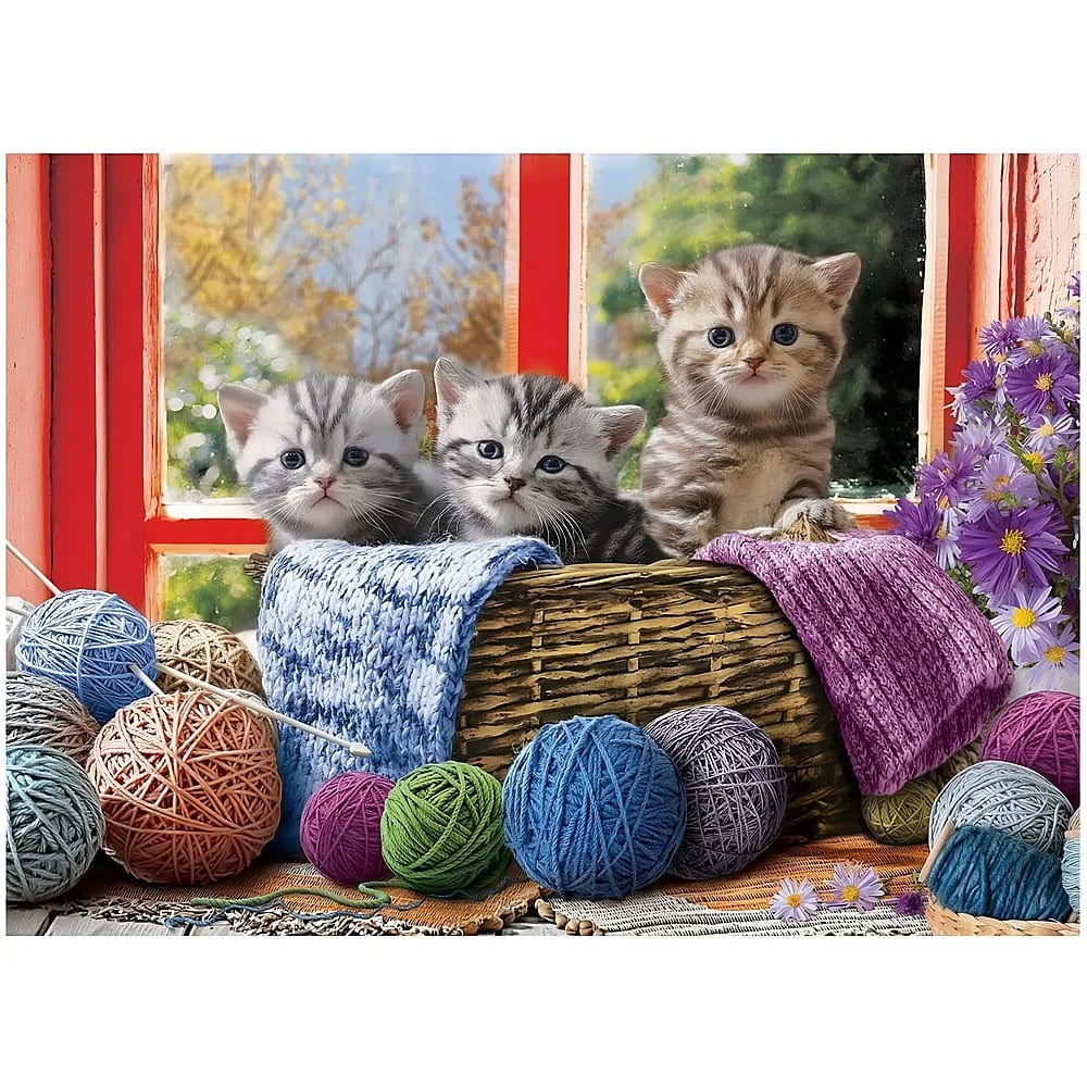 Drei kleine Kätzchen sitzen in einem Korb voller Strickgarn vor einem Fenster mit violetten Blumen.