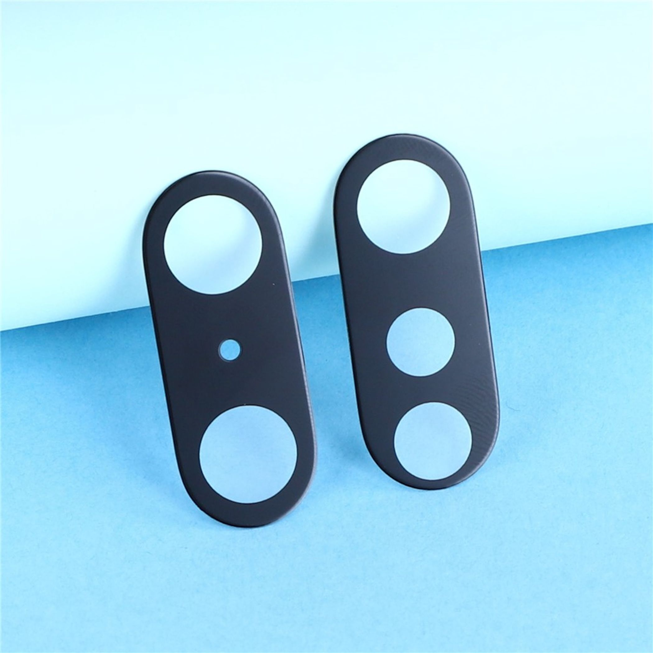 Zwei schwarze Handy-Kamera-Glasabdeckungen mit Aussparungen vor hellblauem Hintergrund.