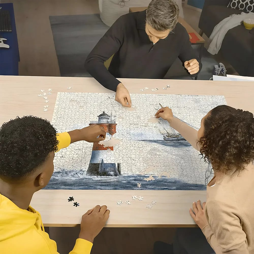 Drei Personen sitzen an einem Holztisch und arbeiten gemeinsam an einem großen Puzzle mit einem Leuchtturm und Schiff.