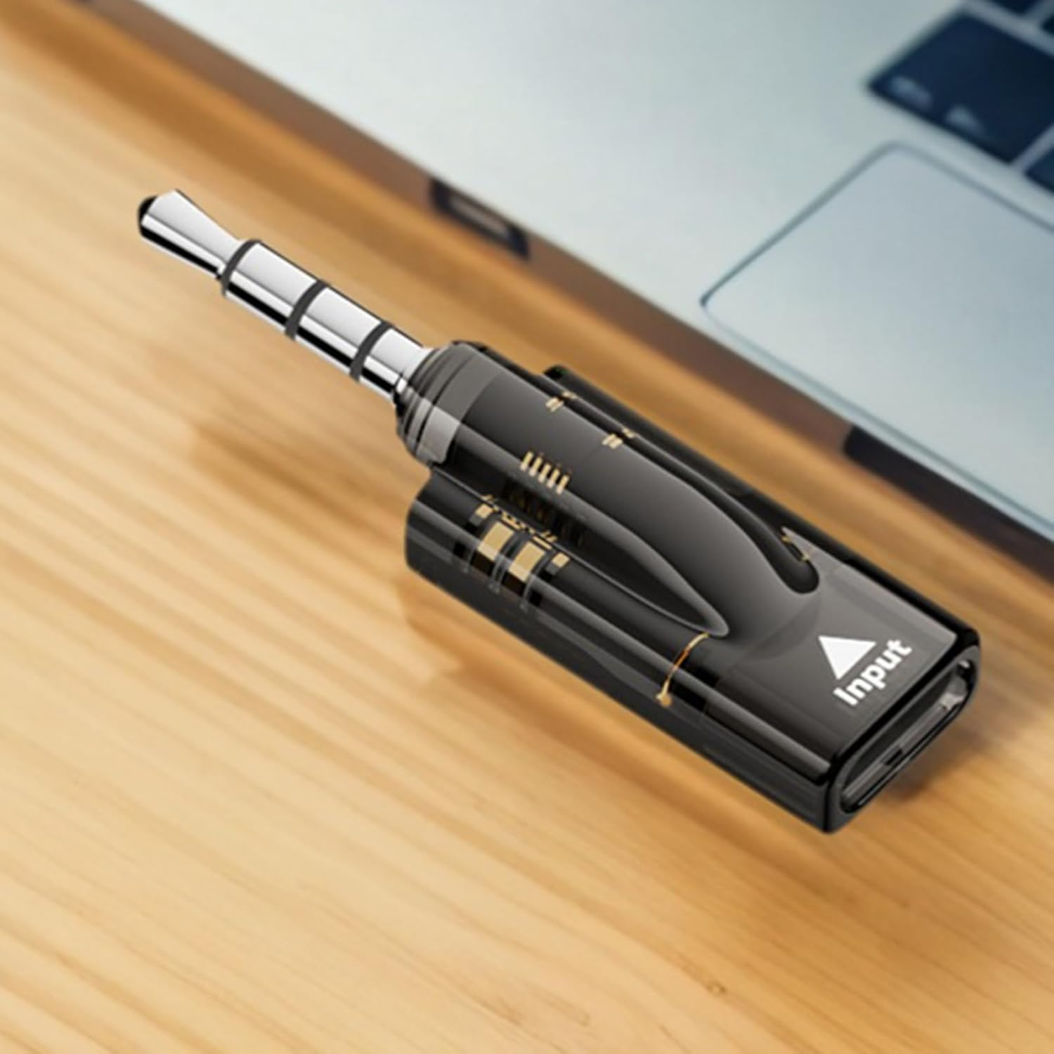 Ein schwarzer Audio-Adapter mit 3,5-mm-Klinkenstecker auf einer Holzoberfläche neben einem Laptop.