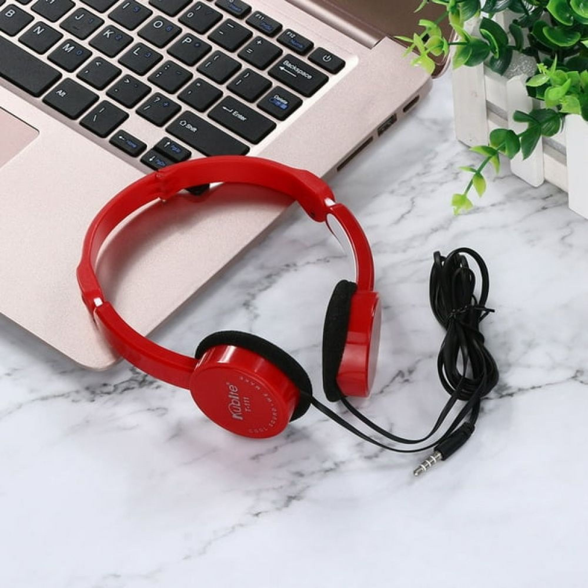 Rote Kopfhörer mit schwarzem Kabel und Klinkenstecker liegen auf weißem Marmor neben einem Laptop.