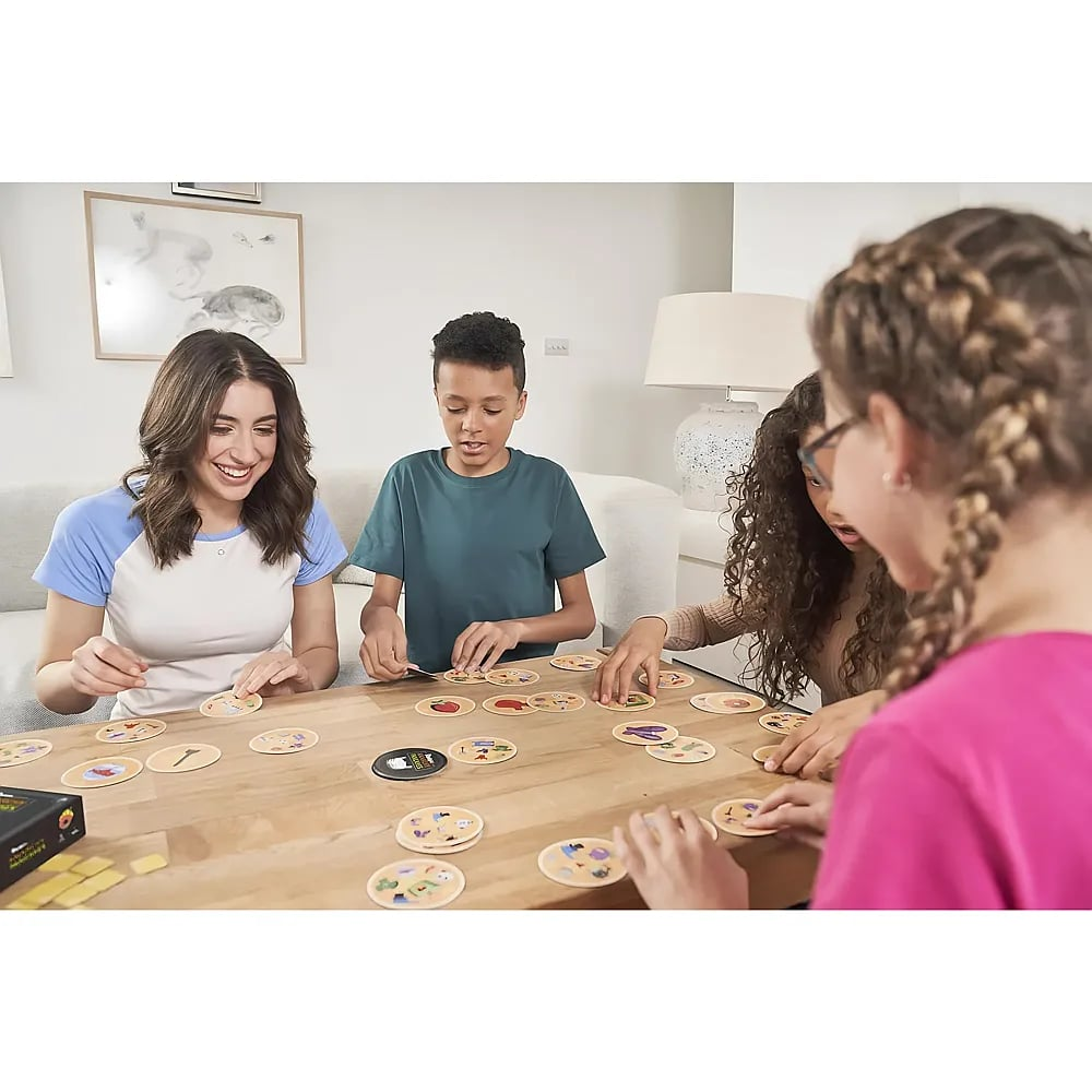 Eine Familie sitzt in einem hellen Raum an einem Holztisch und lächelt, während sie ein passendes Kartenspiel spielt.
