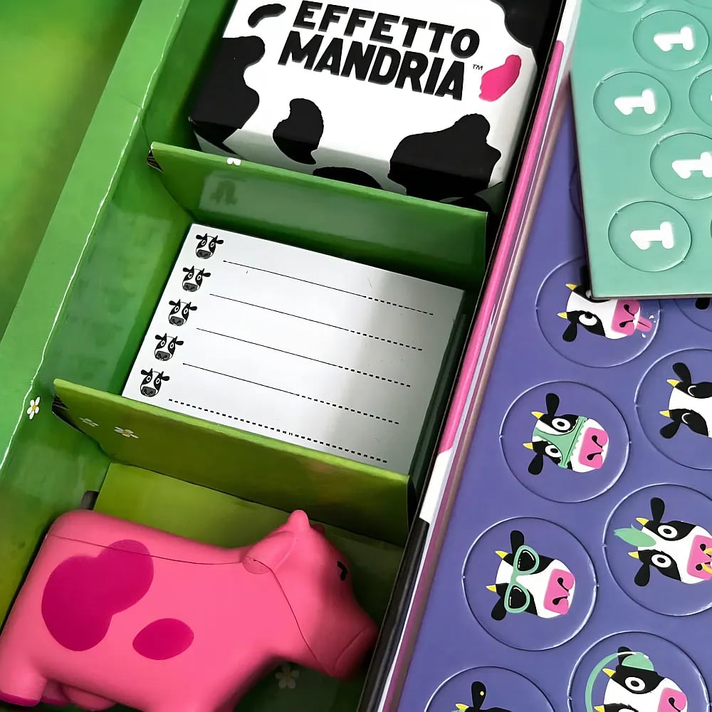 Una scatola di gioco da tavolo a tema mucca con una statuetta di mucca rosa, carte, blocchetti e gettoni.