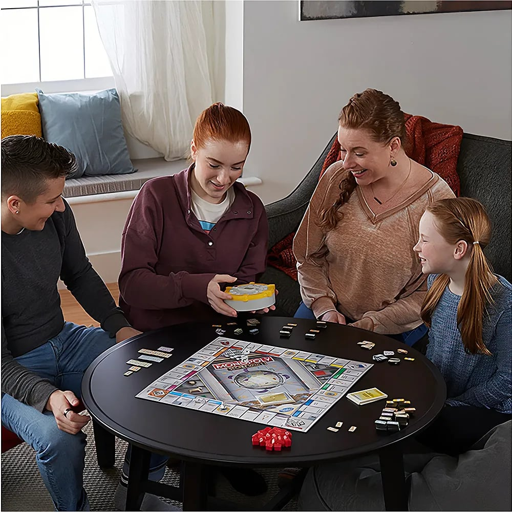 Vier Personen sitzen um einen runden Tisch im Wohnzimmer und spielen vergnügt ein Monopoly-Brettspiel.