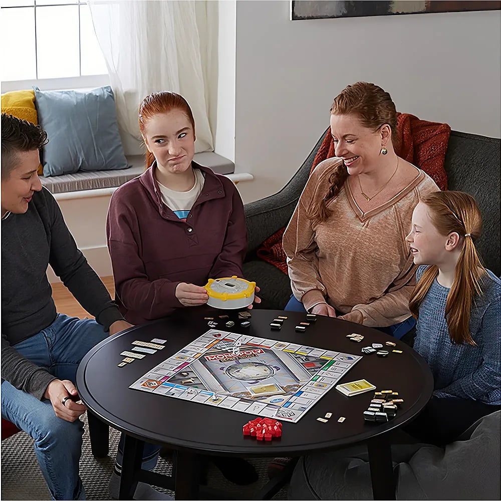Vier Familienmitglieder spielen zusammen Monopoly an einem runden schwarzen Tisch in einem hellen Wohnzimmer.
