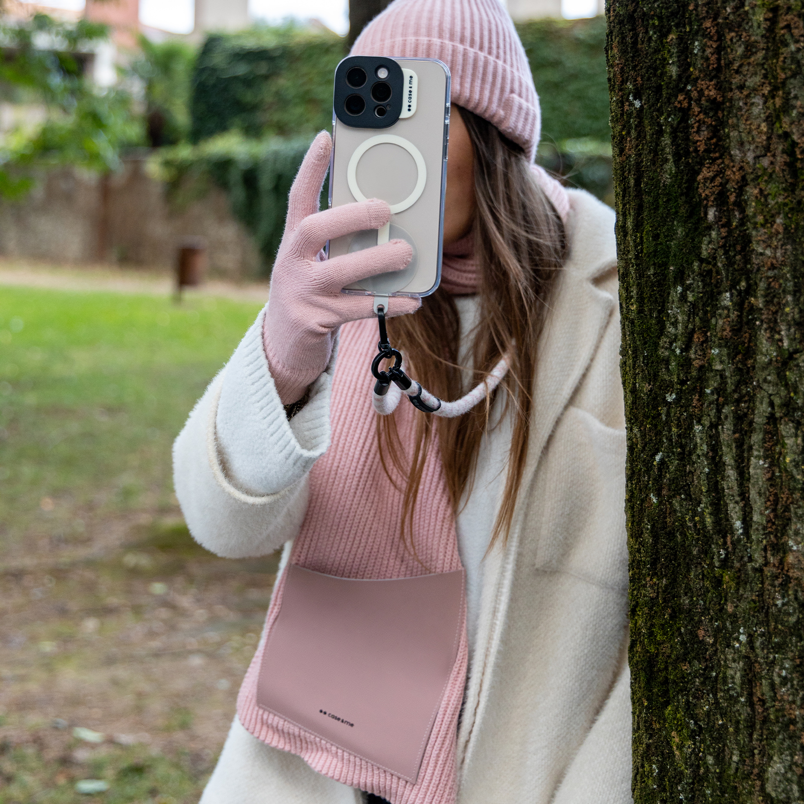 Una mujer con abrigo blanco y accesorios rosados sostiene un móvil con correa en un parque.