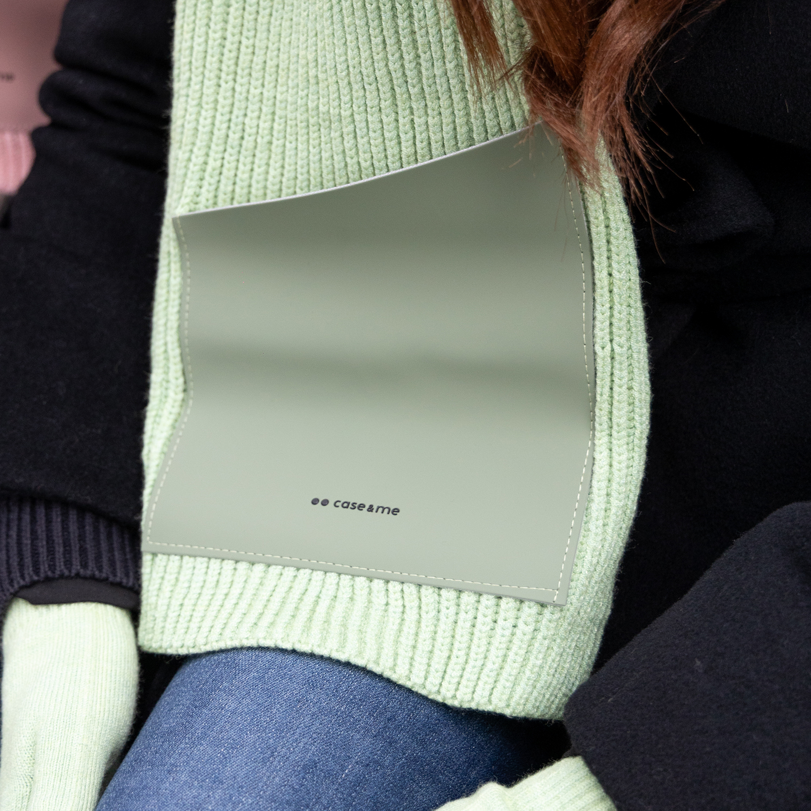 Una funda protectora verde con logo cuelga de una bufanda de punto verde claro, usada con abrigo negro y vaqueros.