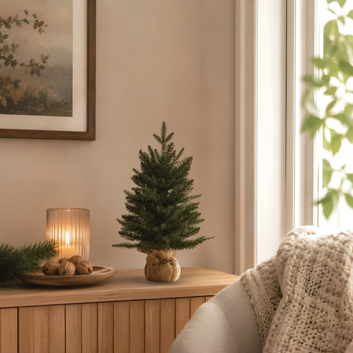 A small Christmas tree in burlap on a wooden table, next to a lit candle and walnuts.