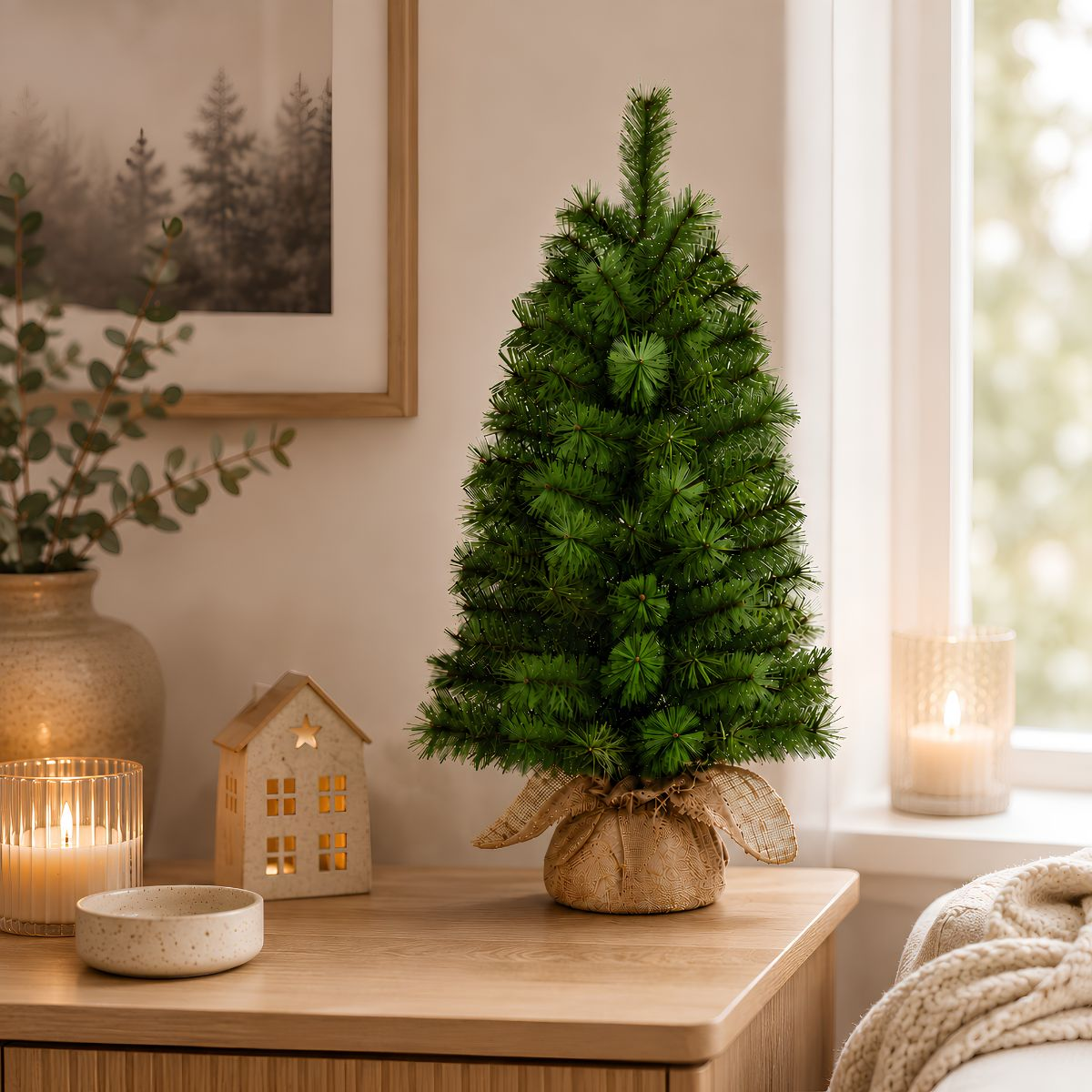 Small Christmas tree in burlap on wooden table with candles and house figurine.