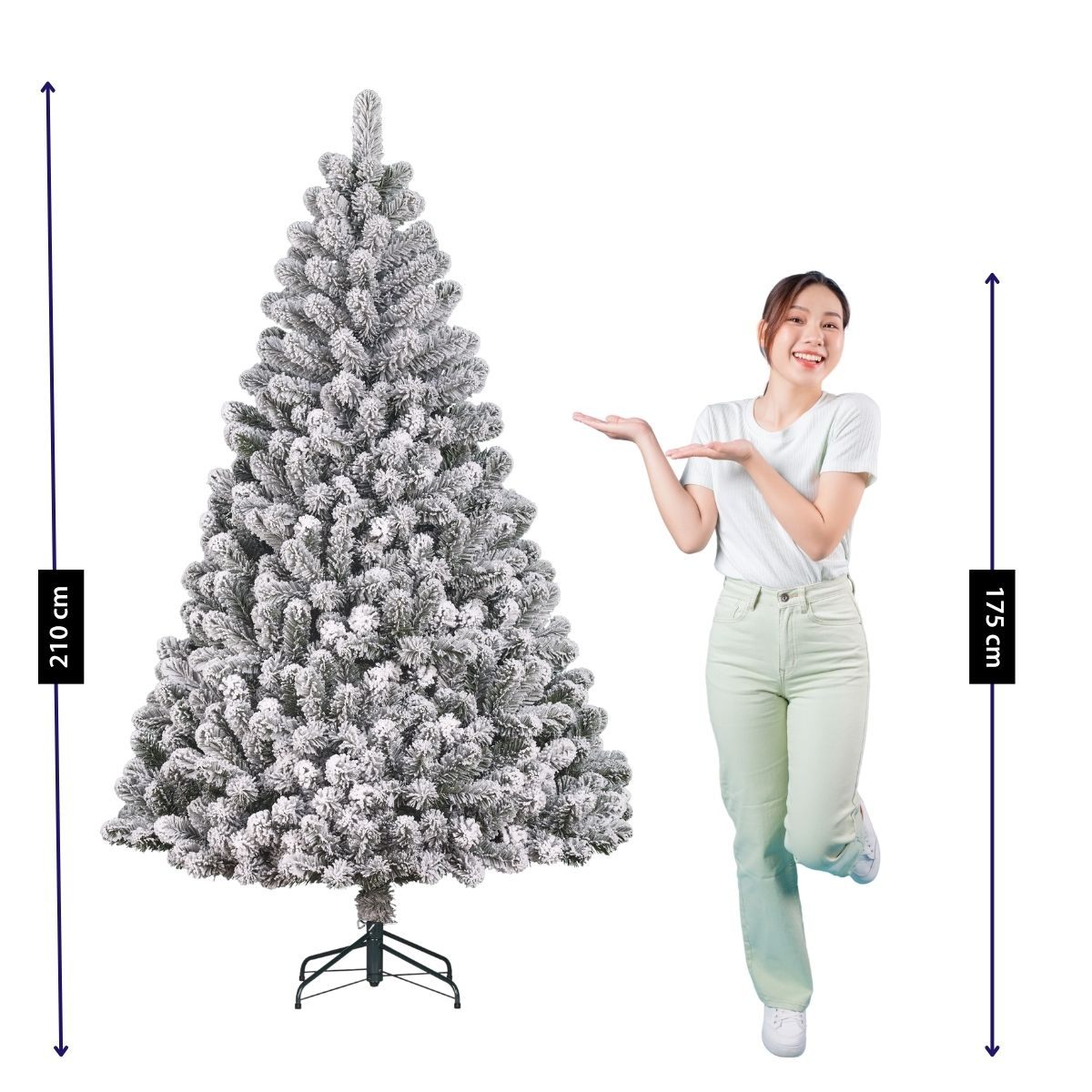 A woman in green clothing gestures toward a large, artificial, snow-covered Christmas tree.