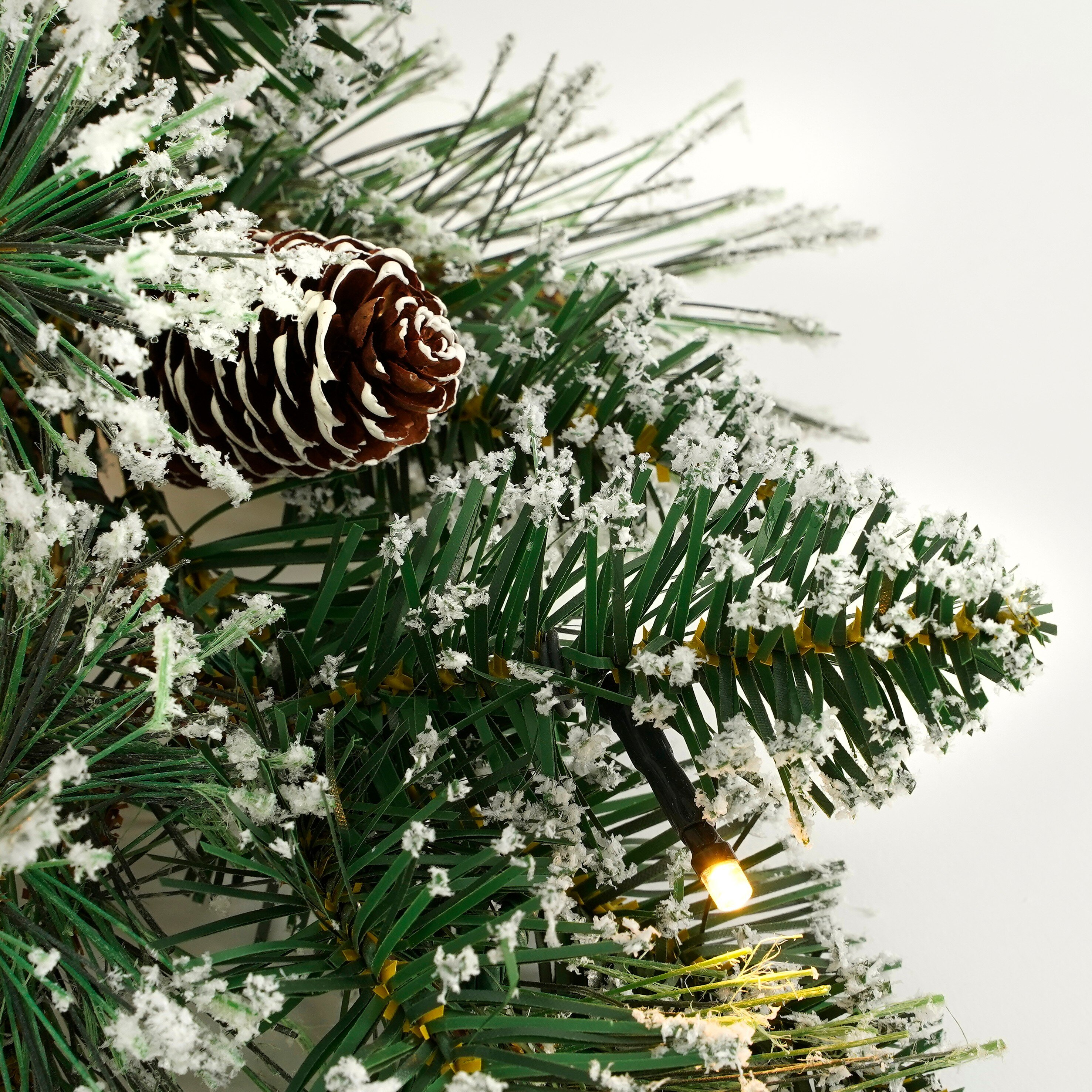 Künstlicher Weihnachtsbaum mit Schnee, Zapfen und kleinen Lichtern auf weißem Grund.