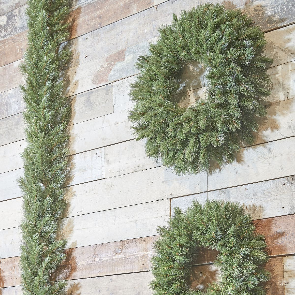 Two green pine garlands and a wreath on a rustic wood plank wall.