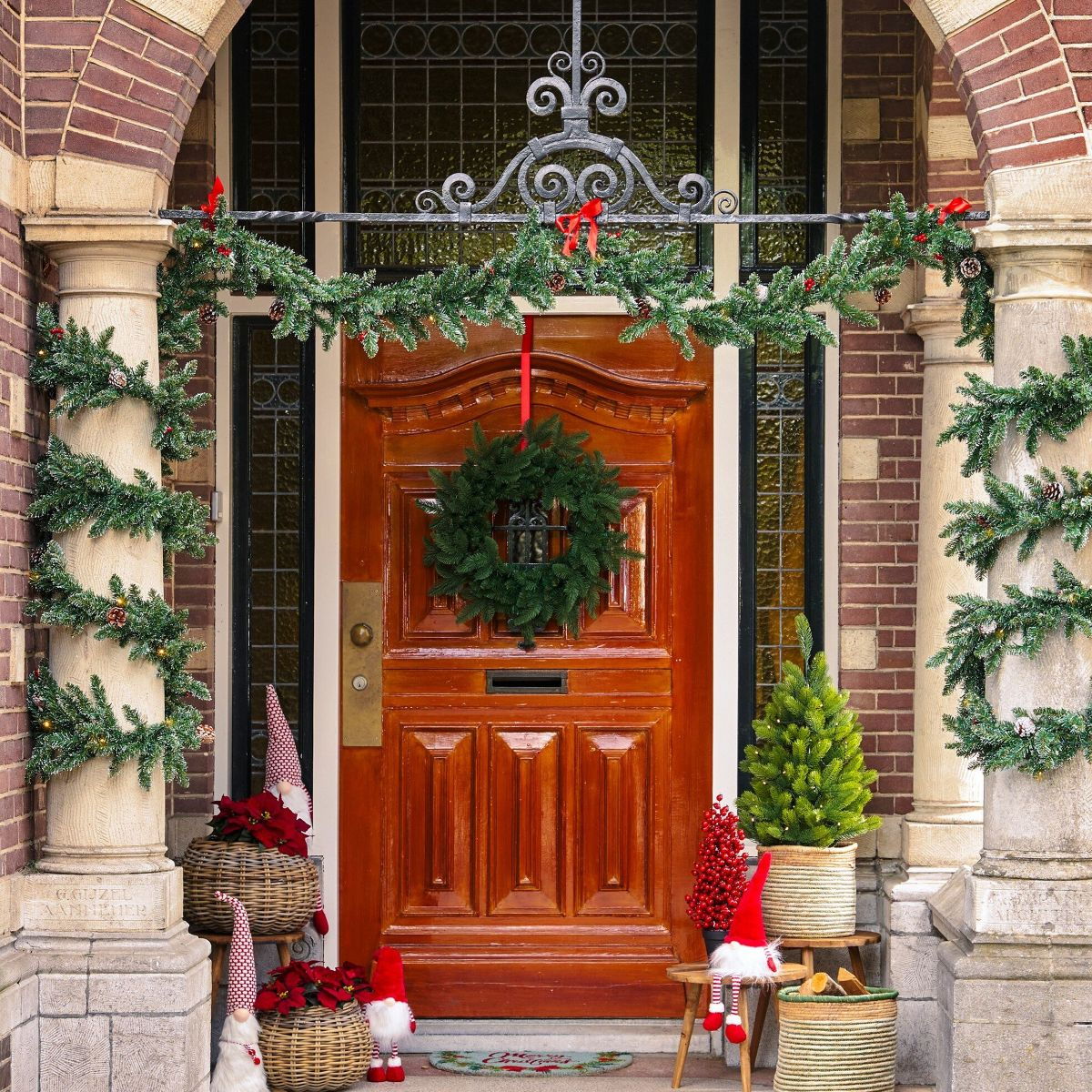 A wooden door decorated for Christmas with a wreath, garlands on columns, small trees, and gnomes.