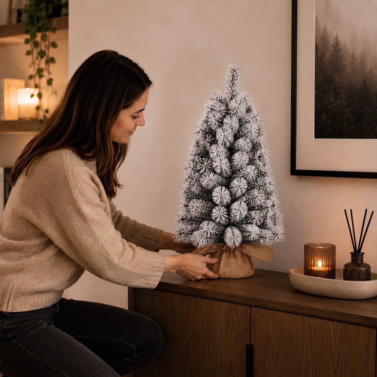 A woman in a beige sweater sets a small, snow-covered artificial Christmas tree on a wooden sideboard.