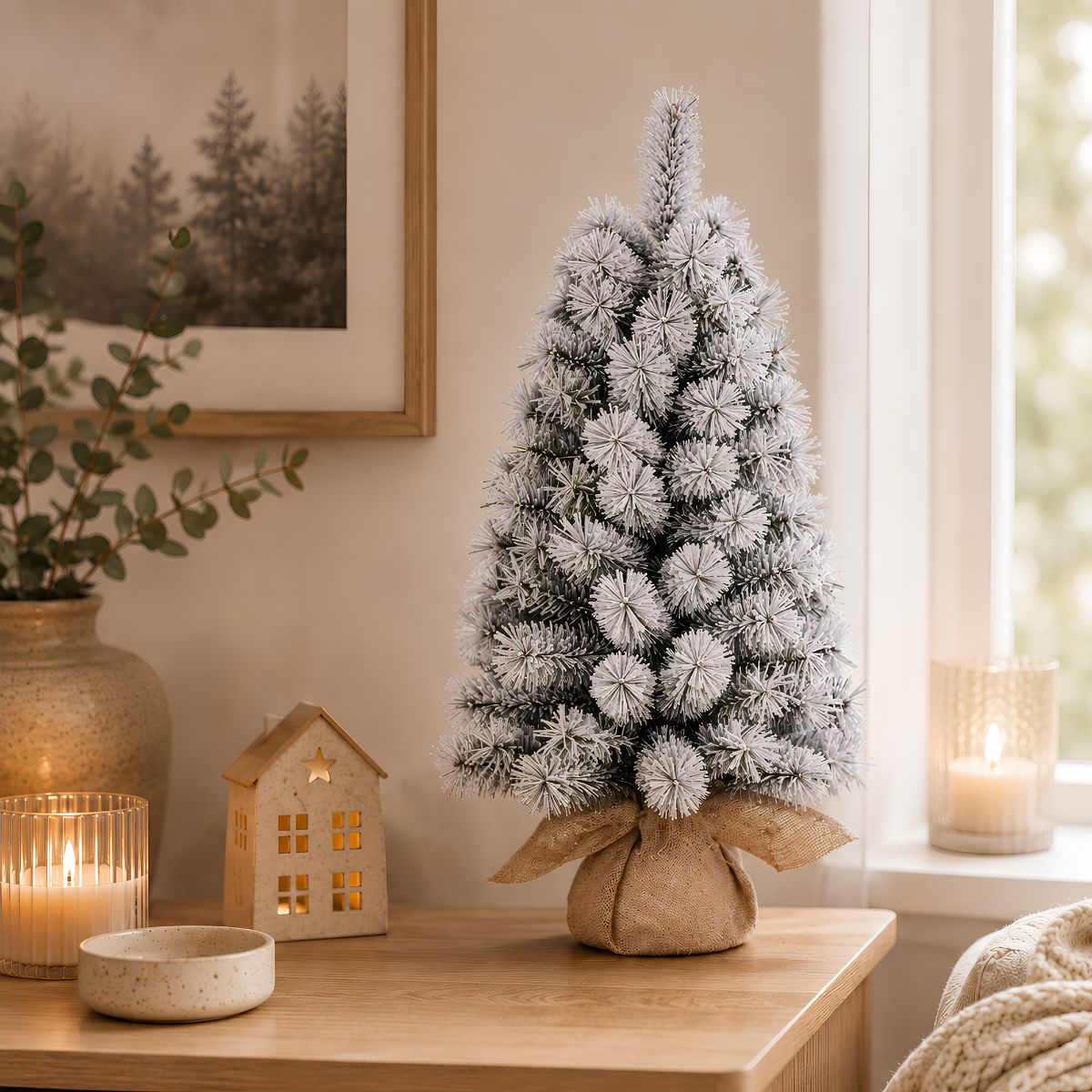 Small snow-flocked Christmas tree on a wooden table with candles, a house-shaped lantern, and eucalyptus.