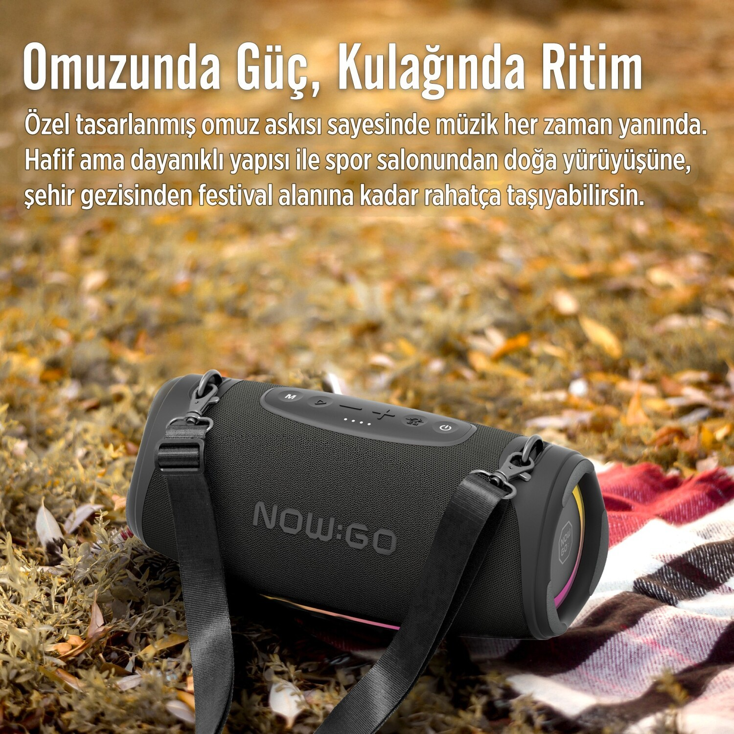 Czarny głośnik Bluetooth NOW:GO z paskiem na ramię leży na kocu w otoczeniu suchych liści.