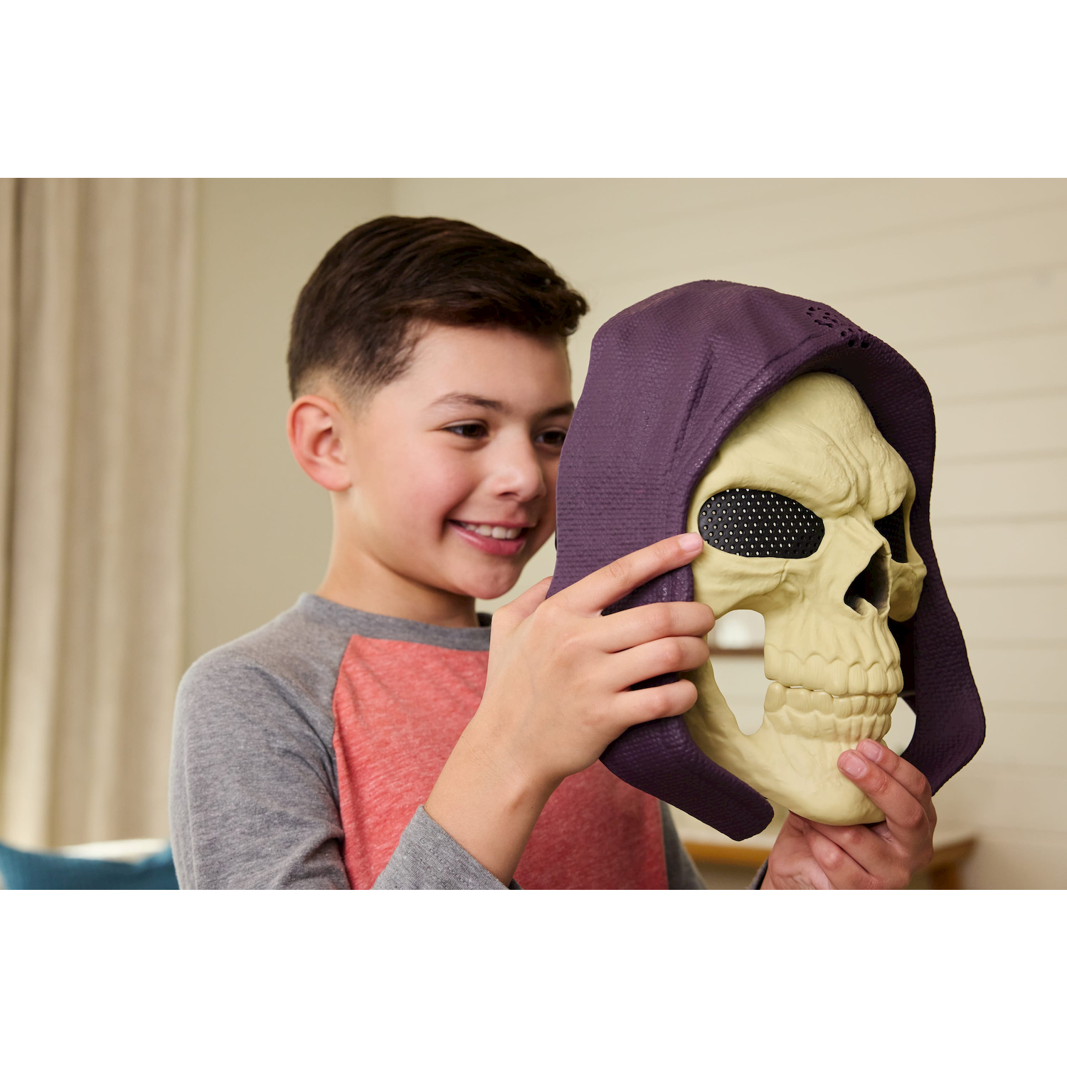 Un niño sonriente sosteniendo una máscara de calavera beige con capucha morada.