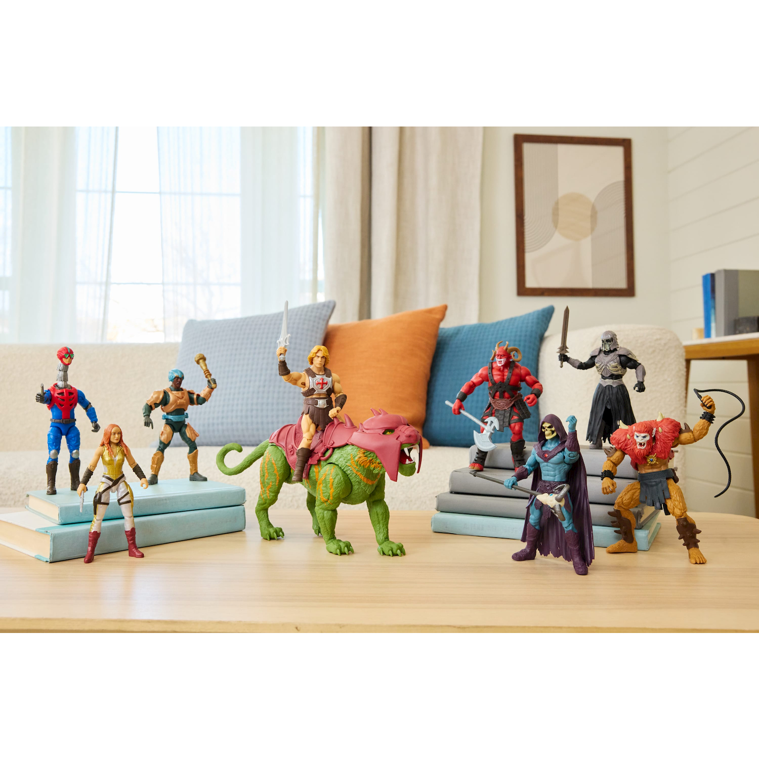 Las figuras de acción de Masters of the Universe están de pie sobre libros en una mesa de madera frente a una sala de estar.