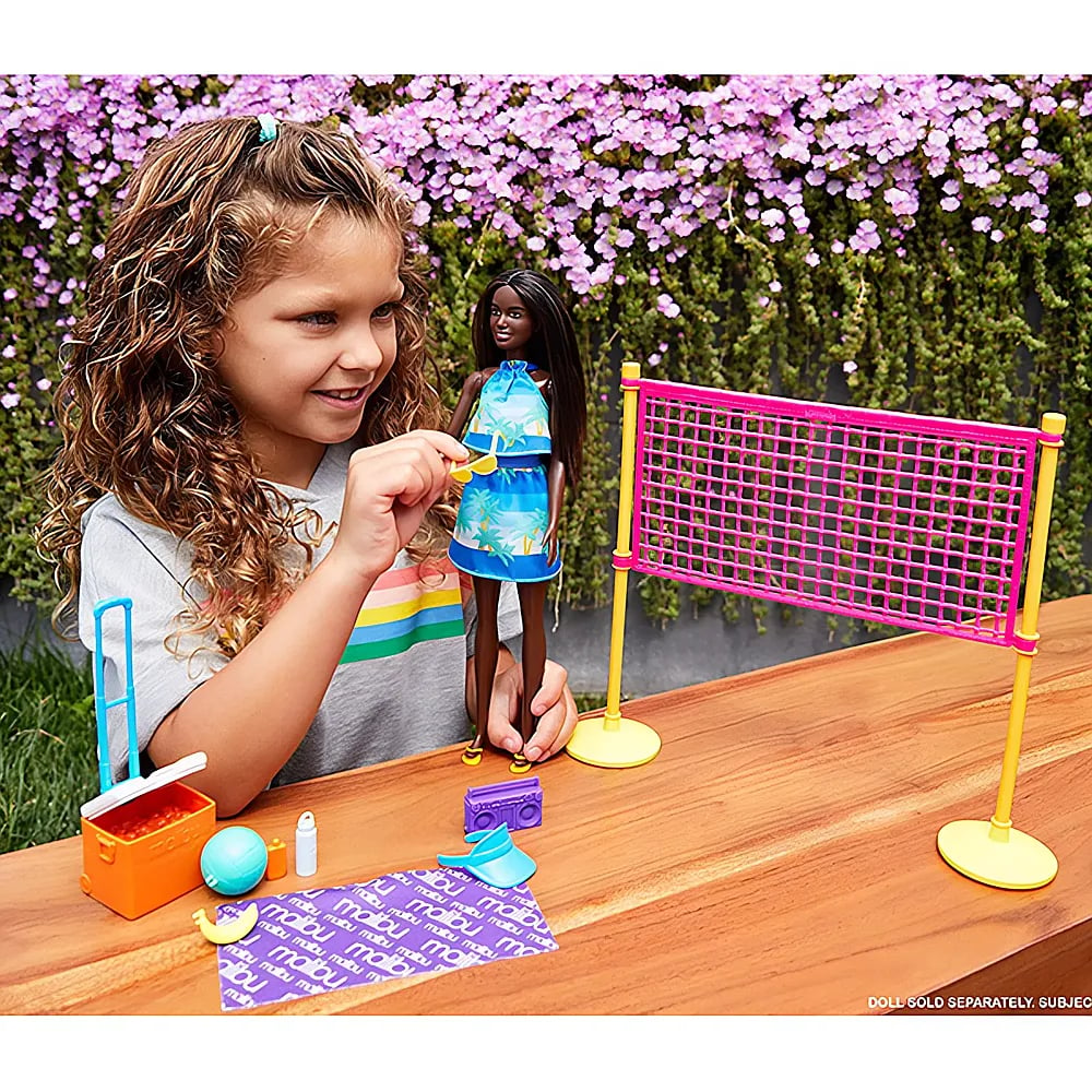 Una bambina sorridente gioca su un tavolo di legno con una bambola, una rete da pallavolo rosa e accessori.