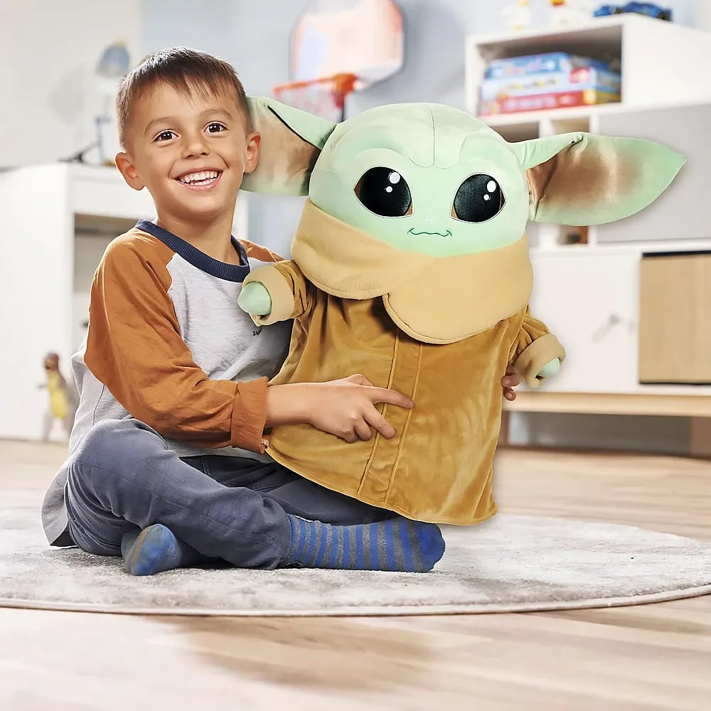 Ein glücklicher Junge sitzt auf einem Teppich und hält ein großes Grogu-Stofftier in einem hellen Kinderzimmer.