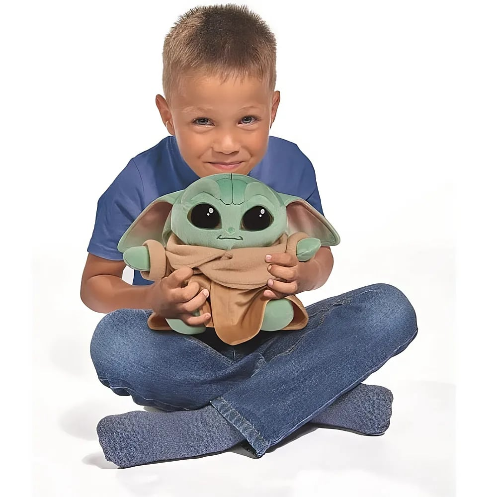 Un giovane ragazzo con camicia blu e jeans siede tenendo un peluche verde di Grogu su sfondo bianco.