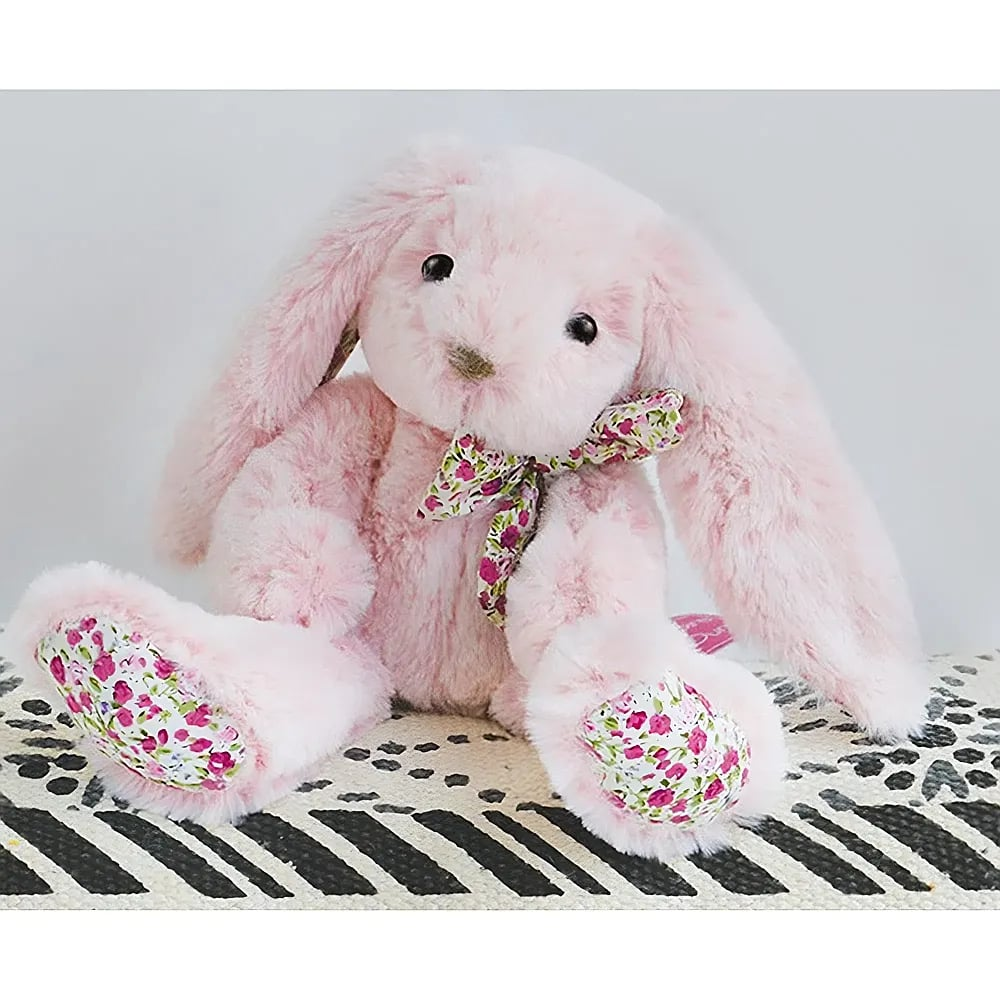 Ein fluffiger rosa Plüschhase mit blumigem Pfotenmuster und passender Schleife sitzt auf gemusterter Oberfläche.