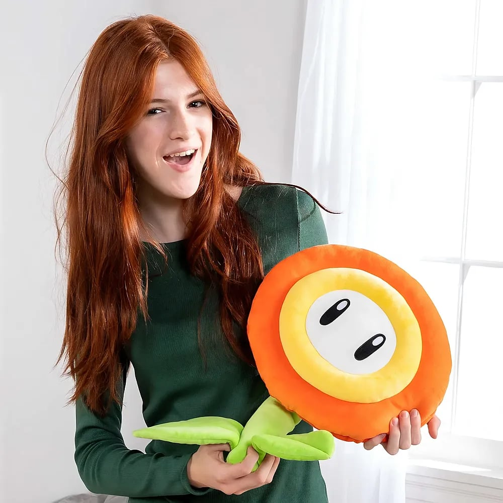 Une femme rousse vêtue d'un haut vert tient une grande peluche en forme de fleur de feu.