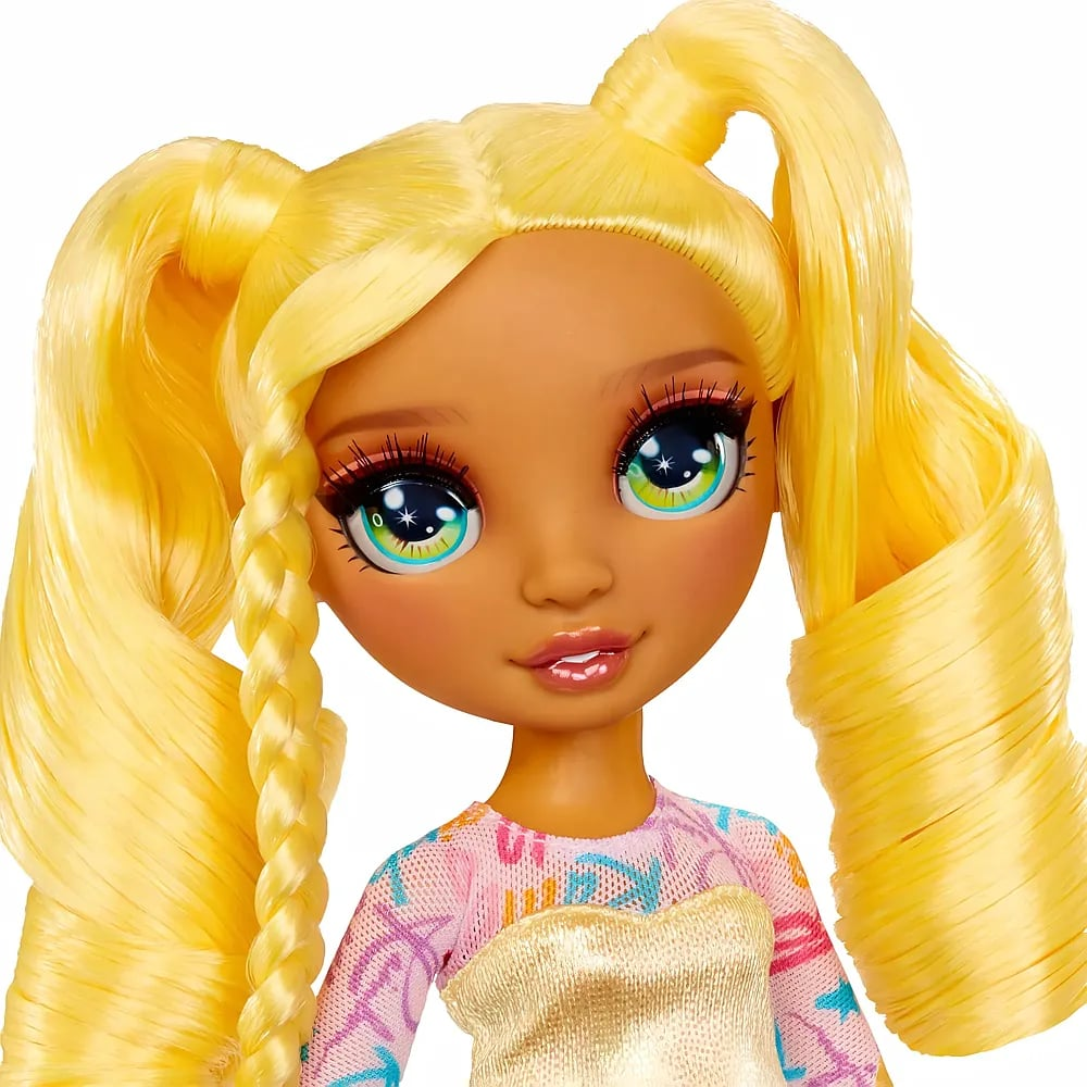 Primer plano de una muñeca con coletas amarillas, grandes ojos azules y top estampado sobre fondo blanco.