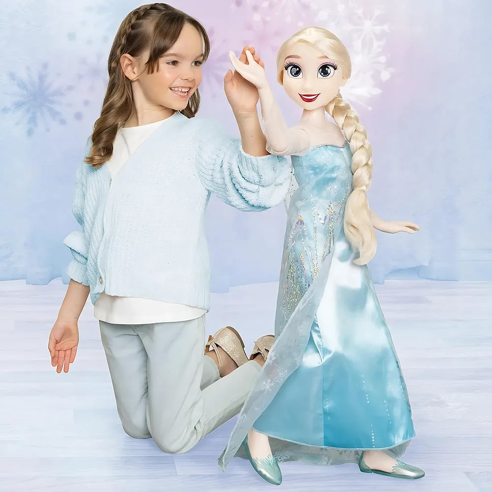Una niña pequeña con ropa azul se toma de la mano con una muñeca grande de Elsa frente a un fondo nevado.