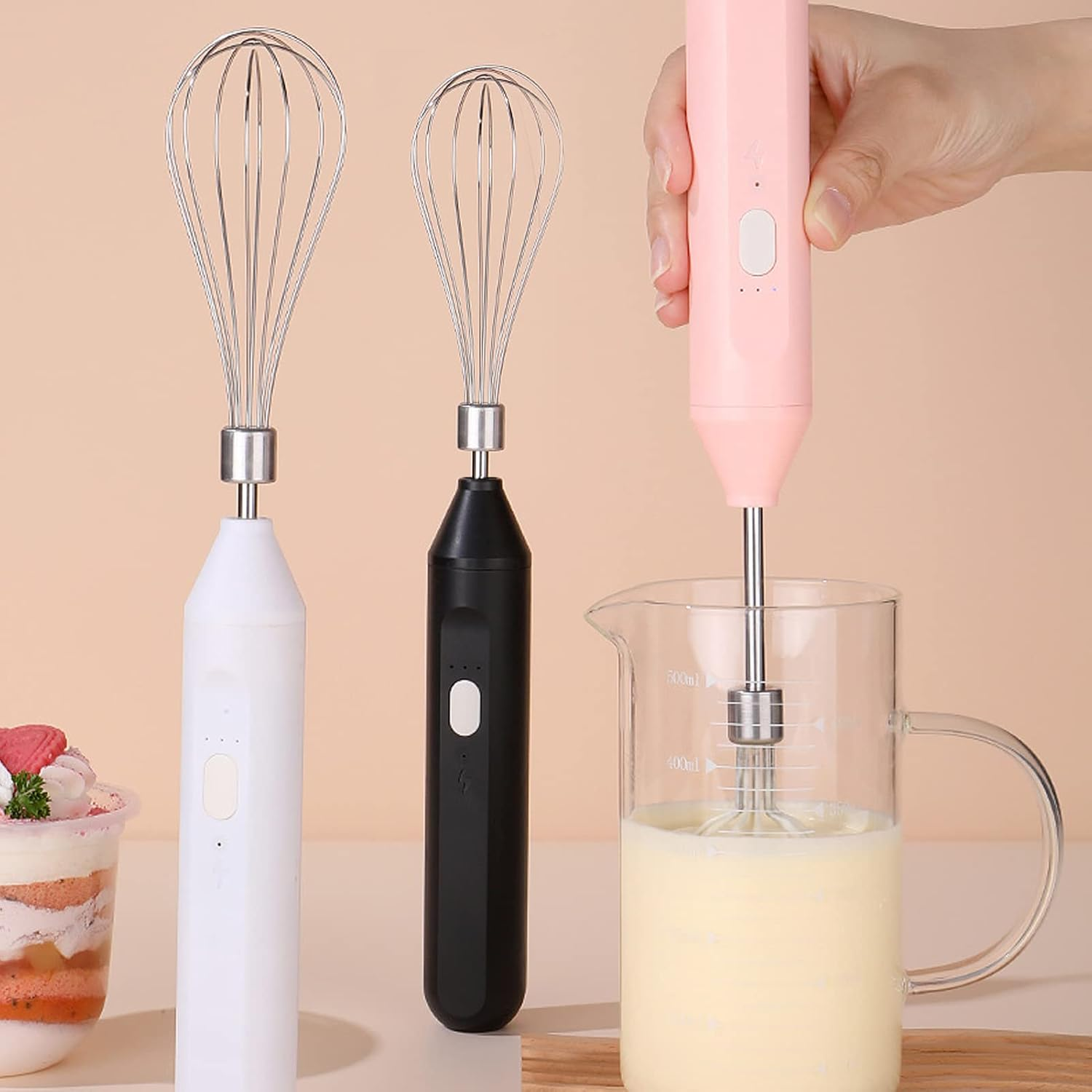 Hand hält einen rosa Handmixer, der Milch in einem Messbecher mixt. Daneben stehen weiße und schwarze Mixer.