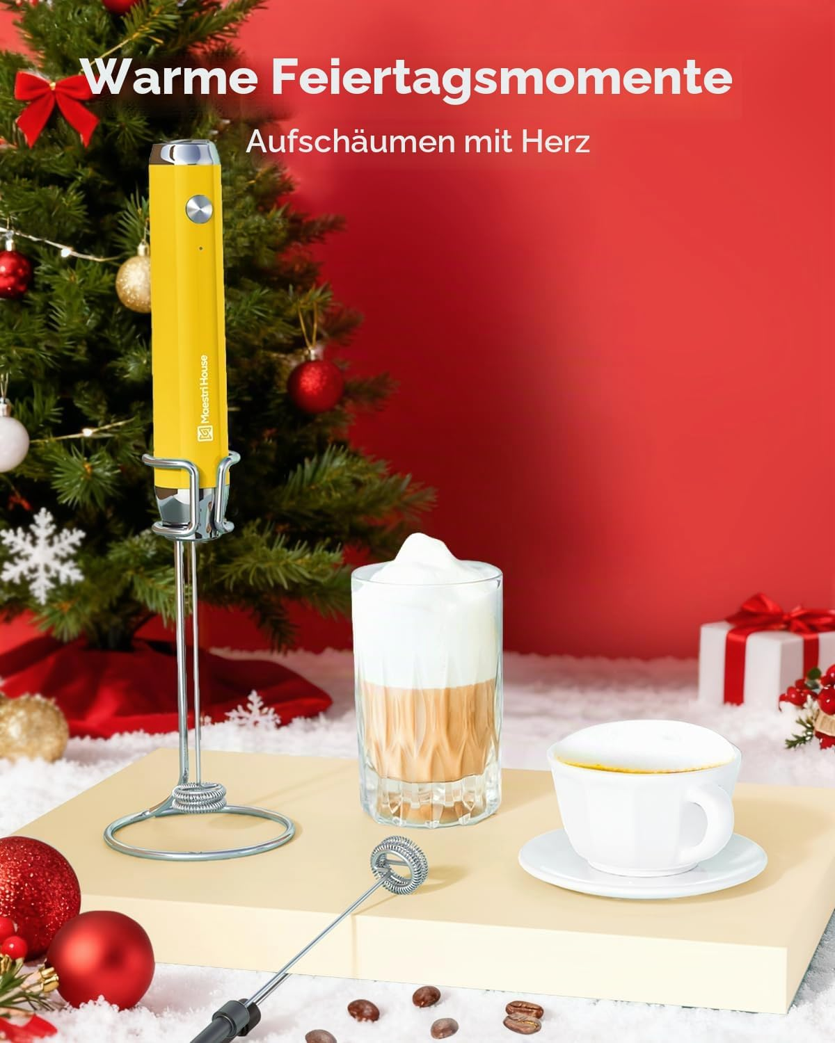 Gelber Milchaufschäumer auf Ständer, Kaffeetasse und Latte-Glas vor einem Weihnachtsbaum mit rotem Hintergrund.