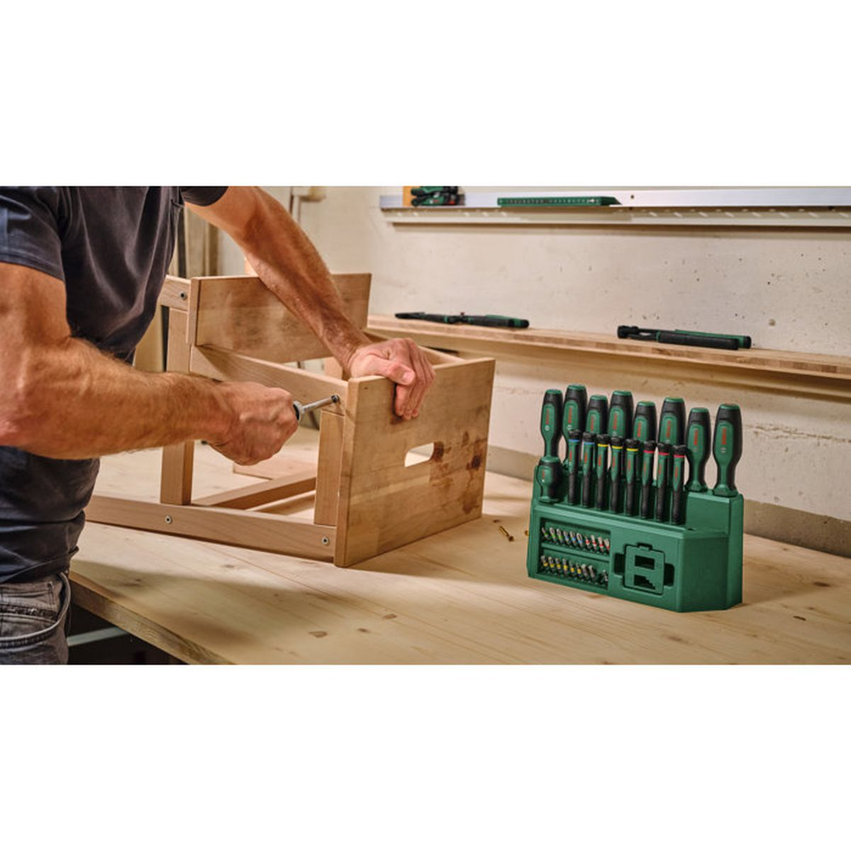 Ein Mann baut einen Holzstuhl an einer Werkbank zusammen, rechts steht ein grüner Bit- und Schraubendreherkasten.
