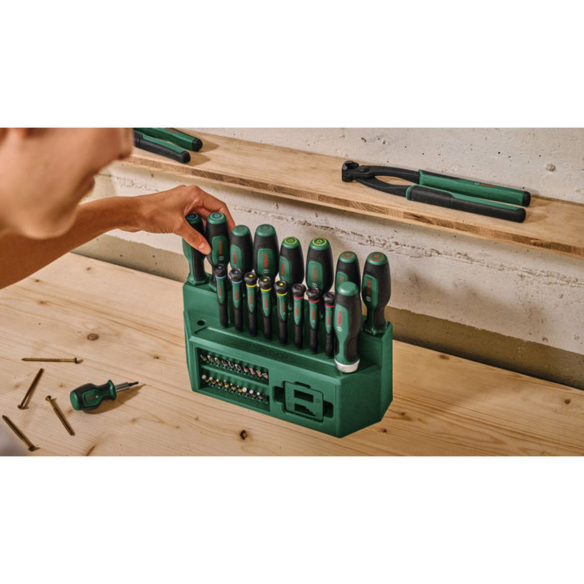 Eine Hand nimmt einen Schraubenzieher aus einem grünen Werkzeugset auf einem Holztisch.