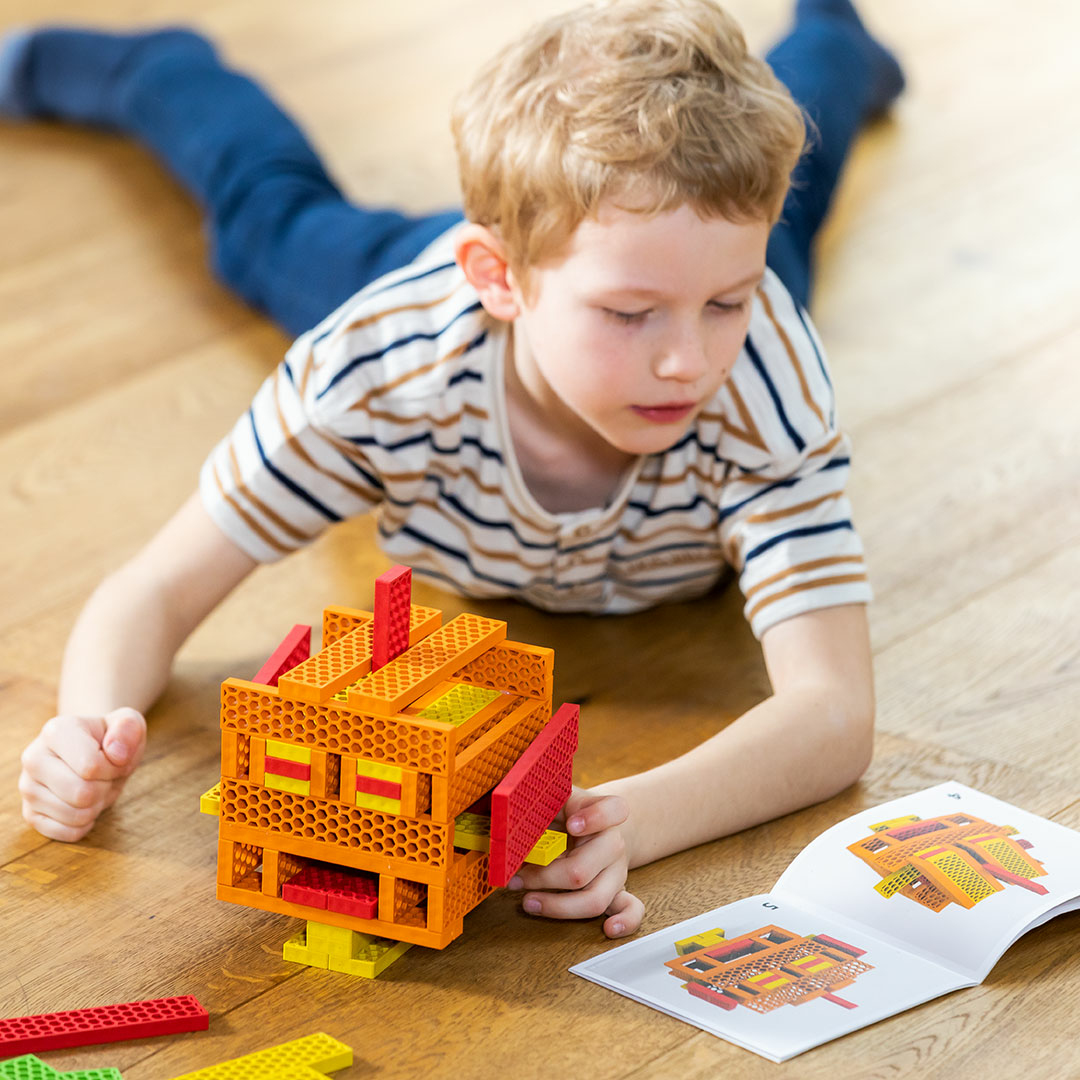 Un bambino biondo sdraiato sul pavimento di legno costruisce un robot con blocchetti colorati.