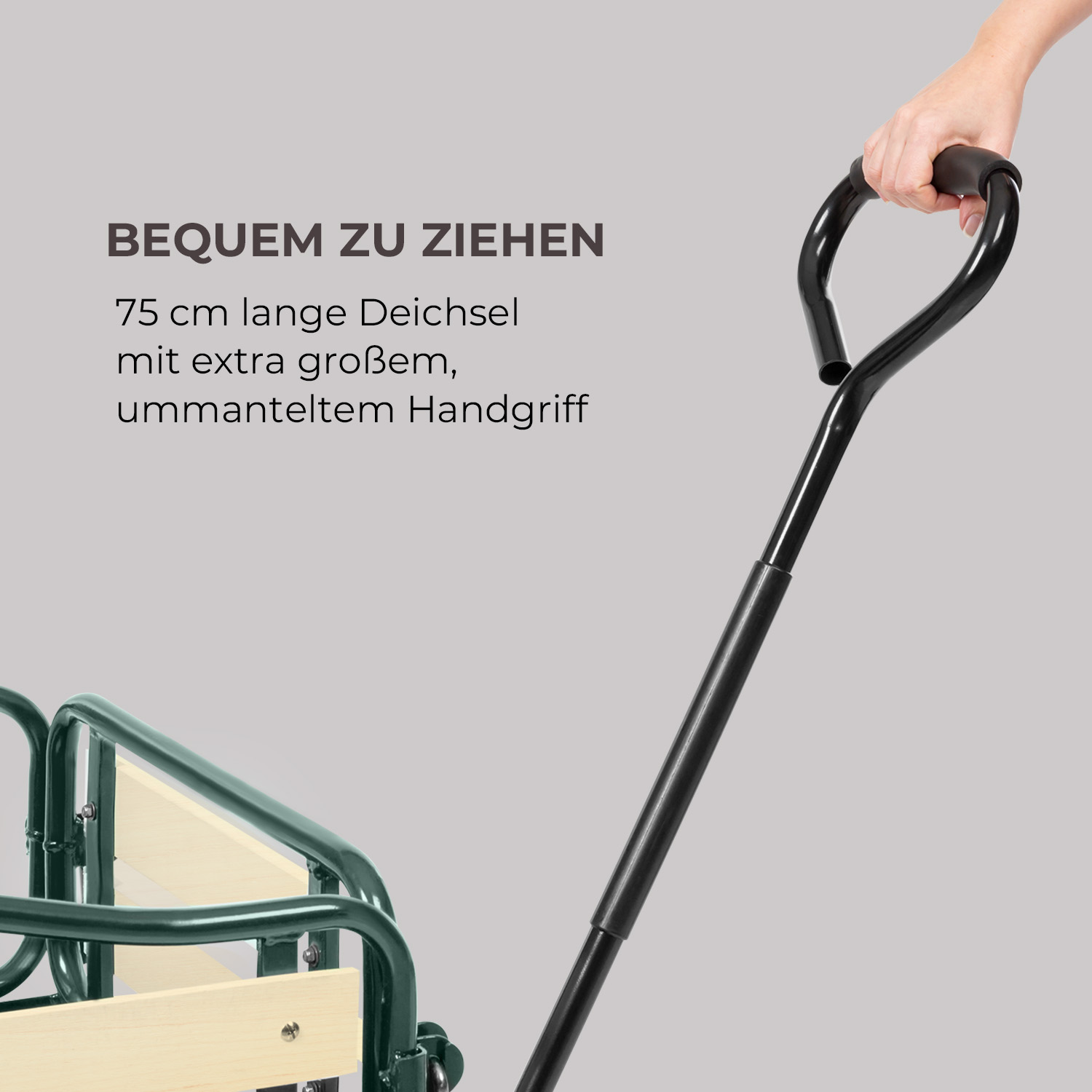 Hand hält die große schwarze Deichsel eines grünen Bollerwagens vor grauem Hintergrund.