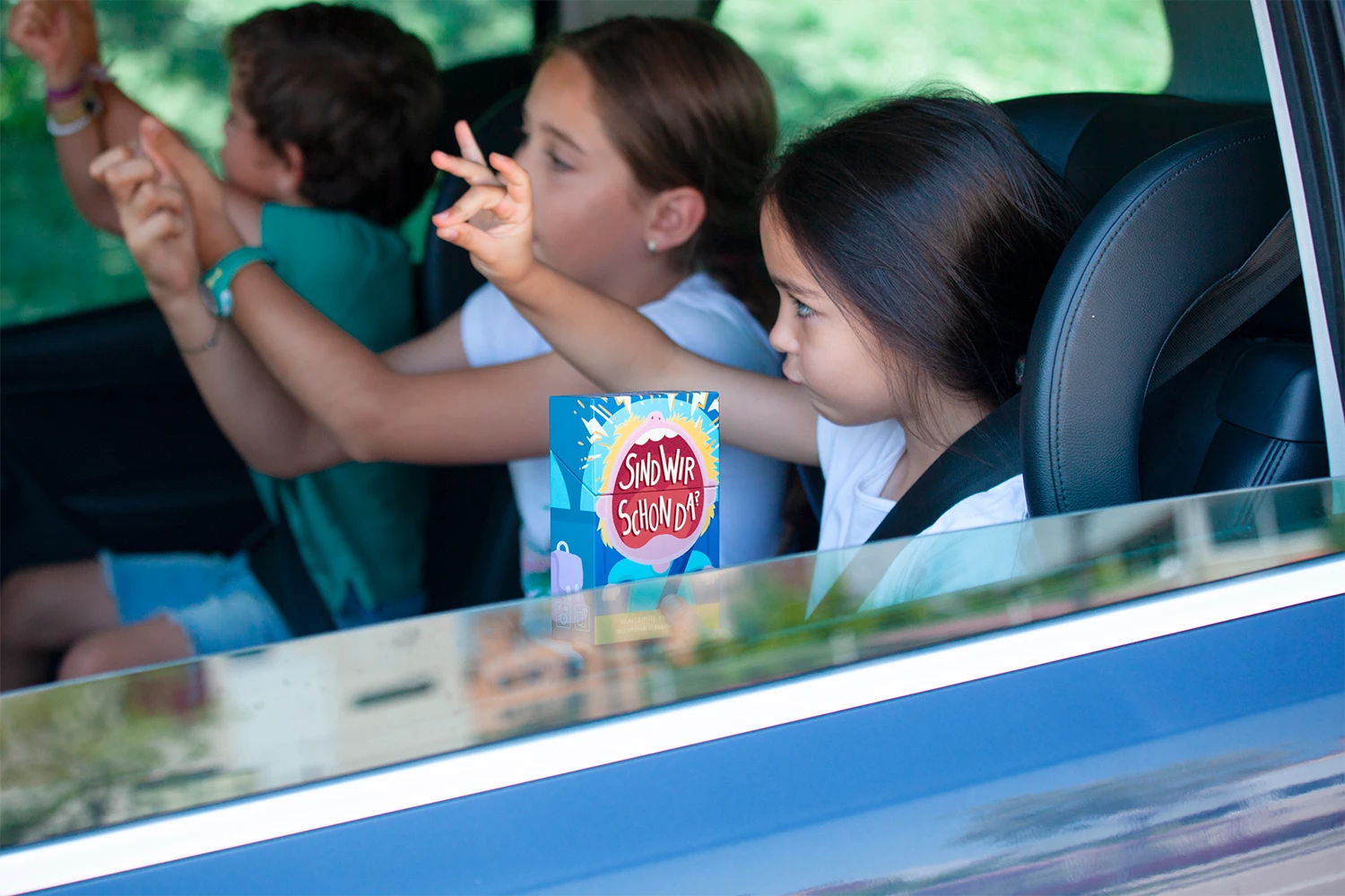 Drei Kinder sitzen im Auto auf der Rückbank und spielen mit einer kleinen Spielschachtel.
