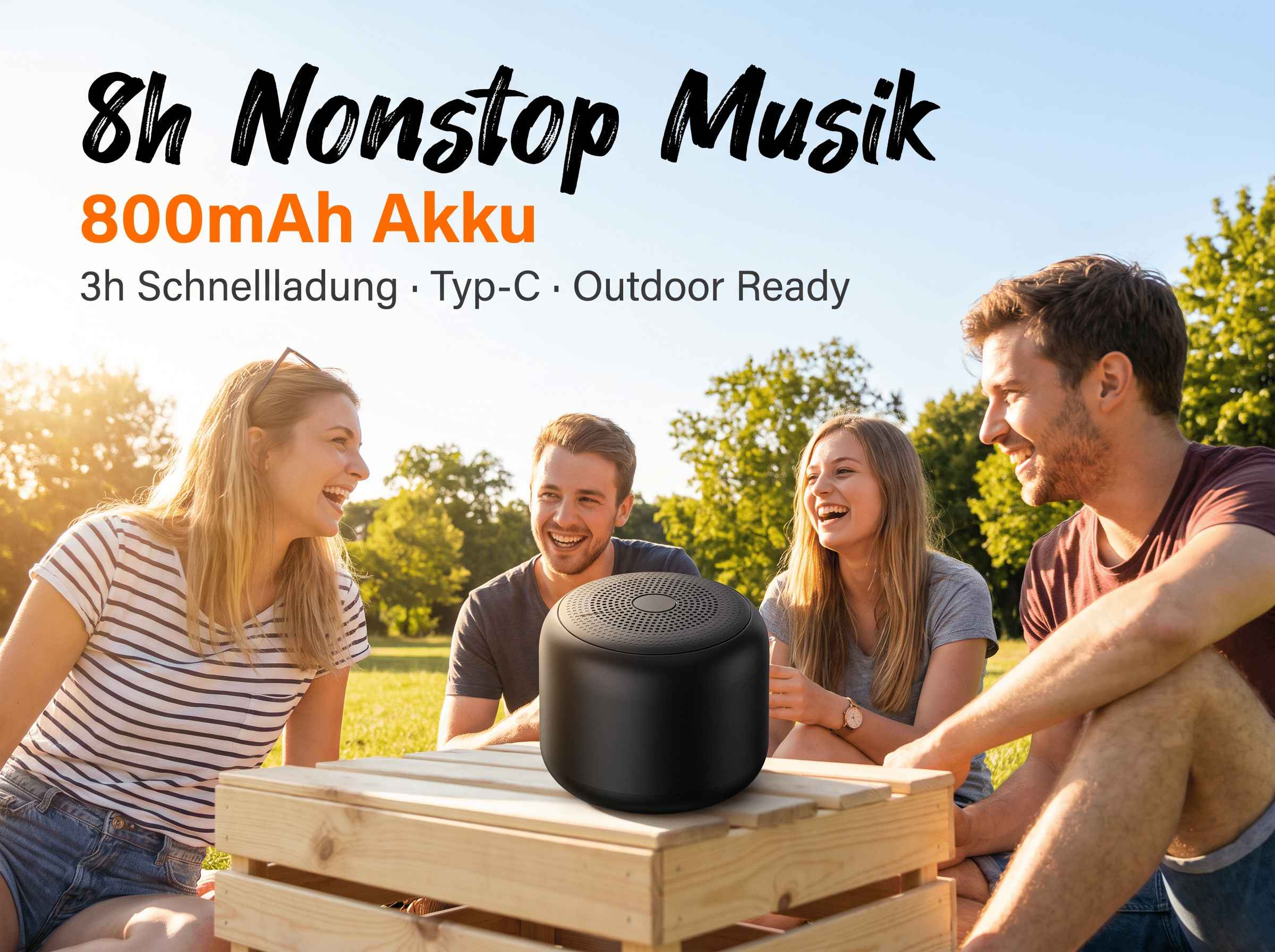Vier glückliche Freunde sitzen draußen auf einer Wiese im Sonnenschein, mit einem schwarzen Lautsprecher auf einem Tisch.