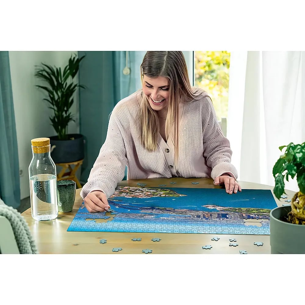 Eine lächelnde blonde Frau sitzt an einem Holztisch und macht begeistert ein großes Puzzle mit einer Küstenstadt.